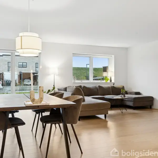 Et spisebord med stole står i en lys stue. Ved siden af er en hjørnesofa. Vinduer viser udsigt til en have og rækkehuse. En reol står i hjørnet.