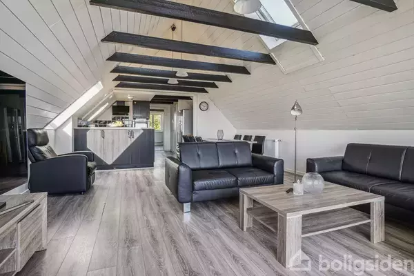Stue med sorte lædersofaer og sofabord på trægulv, under skrå lofter med synlige bjælker. Køkken i baggrunden med spiseplads og ovenlysvinduer, der giver naturligt lysindfald.