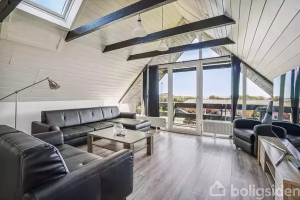 Stue med sorte lædersofaer og sofabord, stående foran store vinduer. Trægulv og skråloft med synlige bjælker tilføjer rummelighed. Udsigten gennem vinduerne viser et grønt landskab.