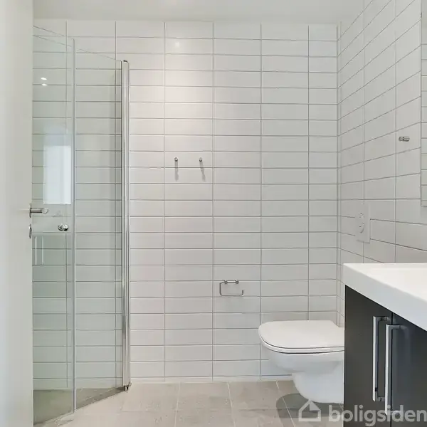 Badeværelse med hvide fliser, brusekabine i glas, væghængt toilet og håndvask med spejl. Enkel, moderne indretning, der skaber et lyst og minimalistisk miljø.
