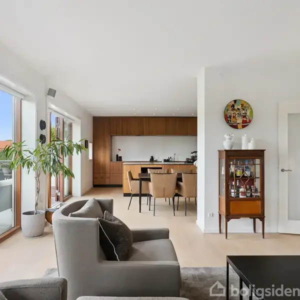 Stue med sofa og sofabord i forgrunden. Spisebord og stole står ved moderne køkken i baggrunden. Store vinduer med udsigt til terrasse og planter.