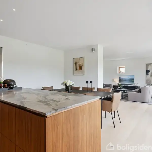 Et moderne køkken-alrum med en stor køkkenø i træ og marmor, omgivet af spisebord og stole. Stuen i baggrunden har en sofa, malerier og store vinduer.