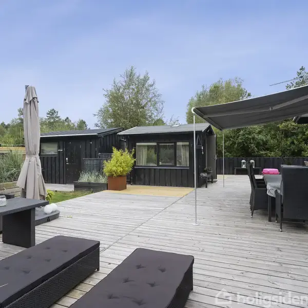 Terrasse med sorte møbler og solsenge, overdækket spisebord, skygget af en stofdug. Omgivet af planter og træer, under en klar, blå himmel.