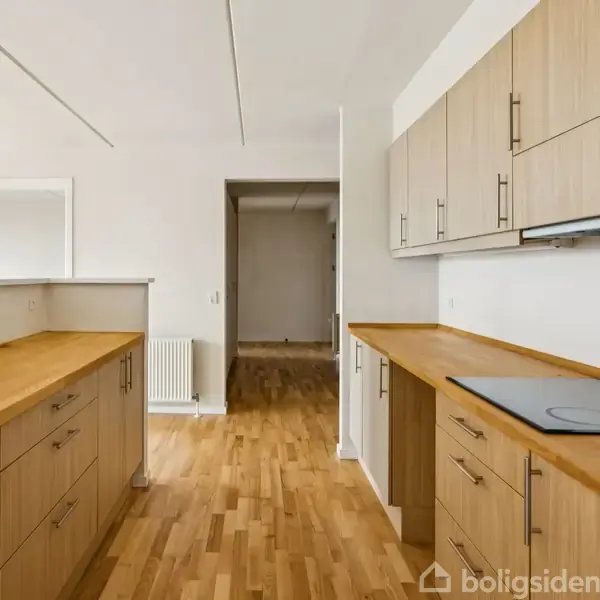 Et moderne køkken med træskabe og -borde. Der er en indbygget kogeplade på bordet. Rummet har lyse trægulve og åbner op til en gang.