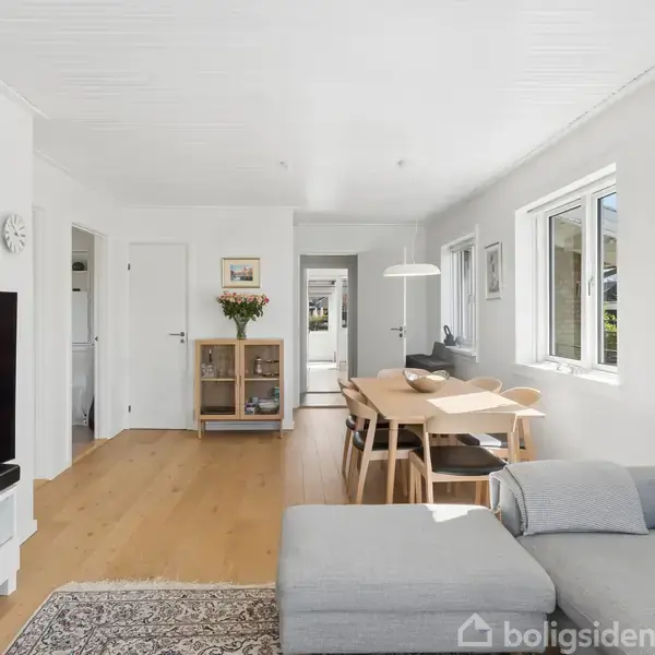 En lys stue med en grå sofa og et fladskærms-tv. Et spisebord med stole står ved et vindue. Træpaneler og lyse vægge skaber en moderne atmosfære.