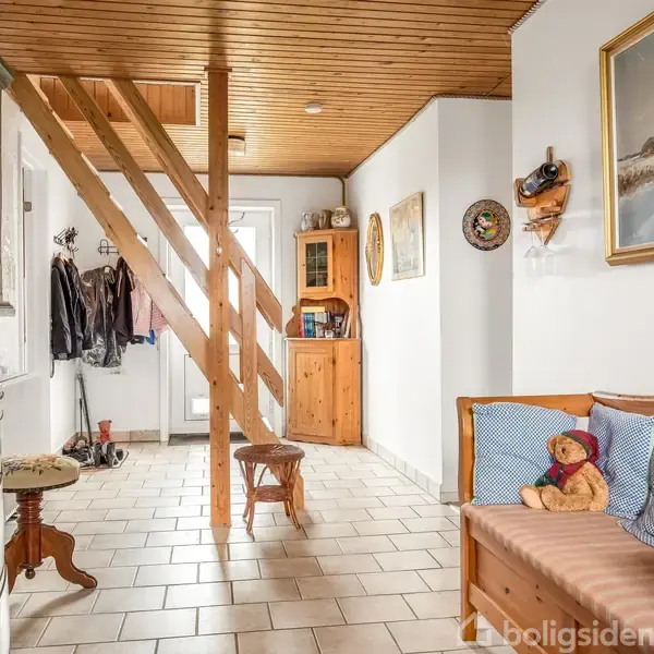 Trætrappe fører til øverste etage, omgivet af hvide vægge og træmøbler. En bruntræsbænk med en bamse står langs væggen; overtøj hænger ved indgangen.