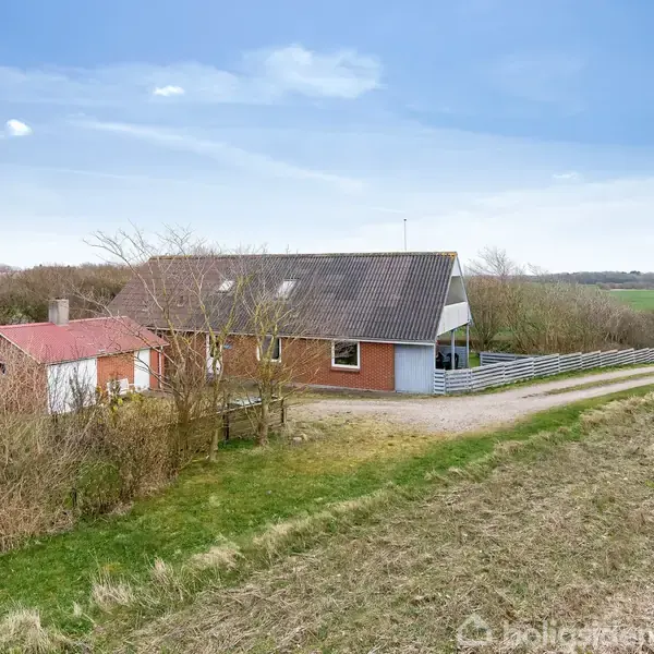 Et rødt murstenshus med mørkt tag står stille omgivet af lav vegetation og grusvej; store, åbne marker strækker sig i baggrunden under en lyseblå himmel uden tekst til stede.
