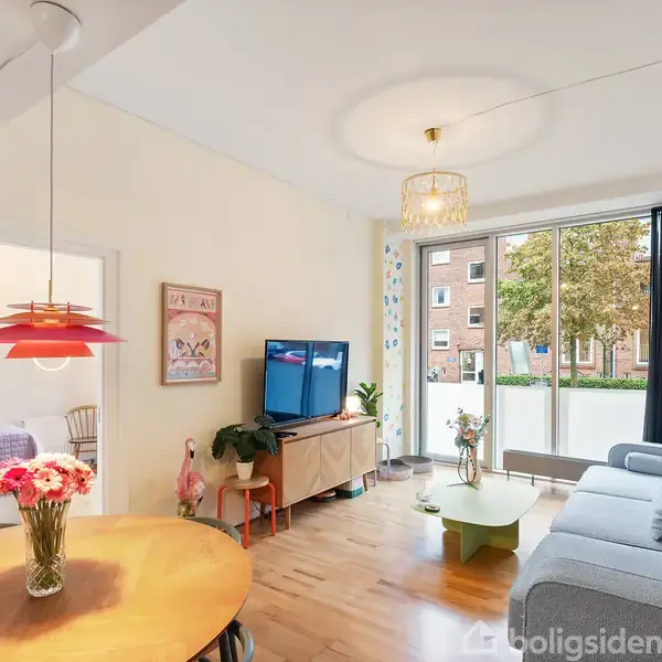 Stue med en grå sofa, et sofabord og et TV på et skab. Rundt spisebord med blomster i vase. Store vinduer giver udsigt til en grøn gård.