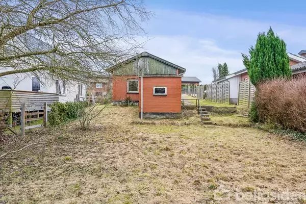 Et rødt hus står i en have med græs omgivet af træer og buske. Ved siden af er der en stentrappe, og yderligere huse kan ses i baggrunden.