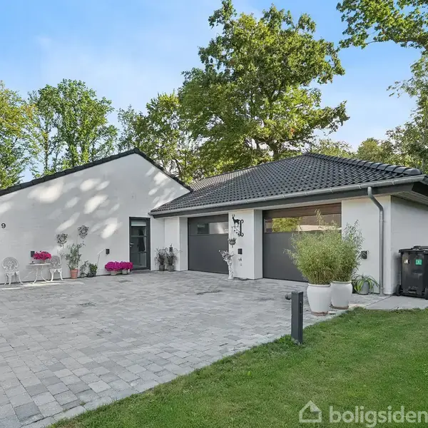 Hvidt hus med gråt tag står ved siden af terrasseomgivelser. Indkørsel er brolagt og dekoreret med blomsterkrukker. Omgivet af frodige træer og græsplæne.