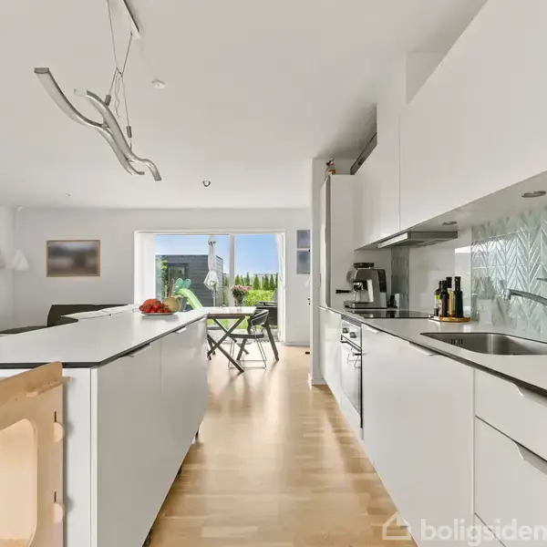 Et moderne, lyst køkken med hvide skabe og en marmor-lignende bagvægge. Køkkenet åbner op til en stue med glasdøre, der fører ud til en terrasse. Trappe til venstre.