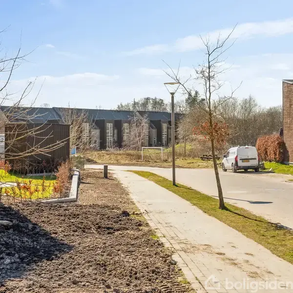Sti snor sig mellem moderne huse og nøgne træer. Parkeret bil ses på vejen til højre. Solrigt vejr med blå himmel.