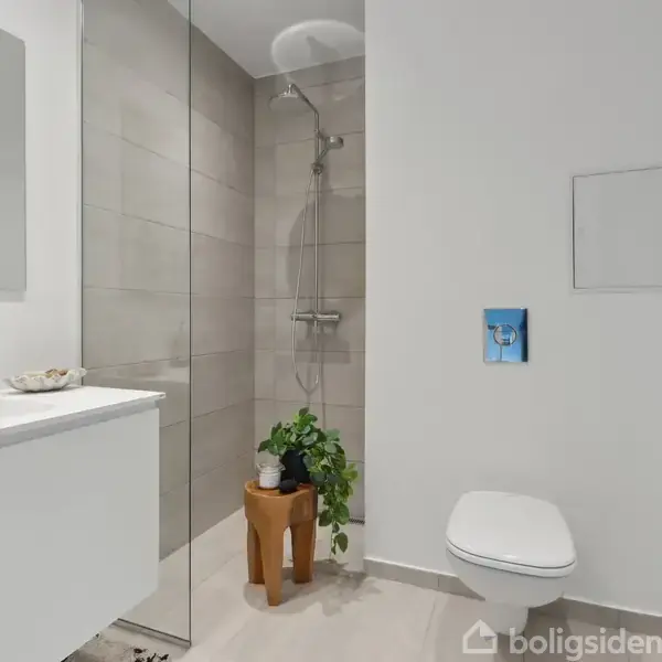 Hvidt badeværelse med bruseniche, vægthængt toilet og håndvask med skab. En grøn plante står på en træskammel ved brusebadet, og der er en stor spejl over vasken.
