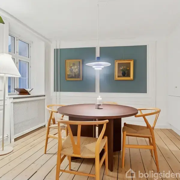Rundt spisebord med fire stole står i et lyst rum med trægulv. En hængelampe hænger over bordet. Væggen har to malerier, og en stor plante ses til venstre.