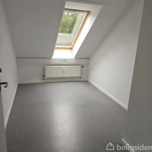 Et tomt rum med et skråt loft har et stort ovenlysvindue, som lukker lys ind. En radiator er monteret på væggen under vinduet.