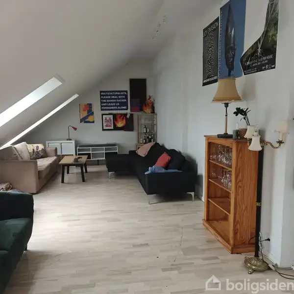 Sofaer står langs en væg i en lys stue med skråvinduer. Et vitrineskab holder en lampe. Plakater hænger på væggen. Tekst: “RELAXOPHOBIALETICS, PLEASE DEAL WITH THE DANGER MACHINE AND ALWAYS LET THE FORBIDDEN ALONE.”