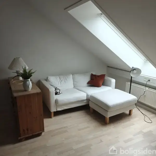 En hvid sofa med puder står i et lyst rum under et skråvindue. Ved siden af er en trækommode med lampe og plante. En gulvlampe står ved radiatoren.