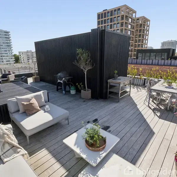 Tagterrasse med moderne møblering, herunder grå sofaer og borde med planter. Grønne planter og blomster omkranser området. Bygninger i forskellige stilarter omkranser terrassen under en blå himmel.