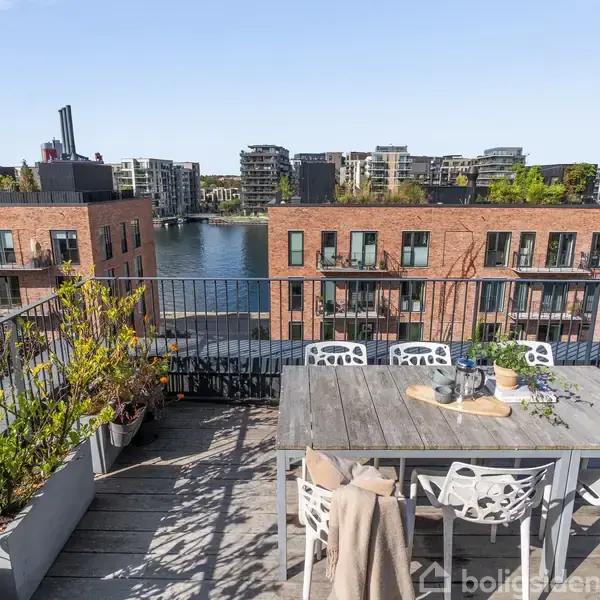 Bord med stole står på en tagterrasse med udsigt til en kanal. Omkranset af moderne røde murstensbygninger med flere altaner og store vinduer. Plantekasser pynter langs rækværket.