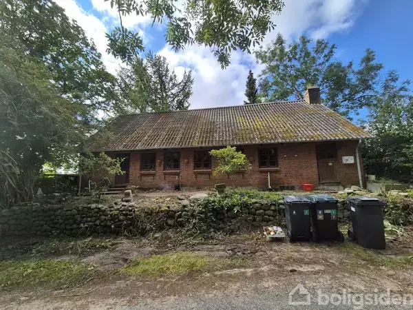 Et gammelt rødt murstenshus står på en grøn grund omgivet af træer; forhaven har affaldscontainere foran en lav stenmur.