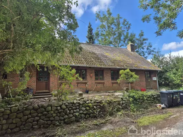 Et murstenshus med mosdækket tag står omgivet af frodig vegetation og stenmur. Der er affaldscontainere i højre forgrund. Skyfri himmel ses over stedet.