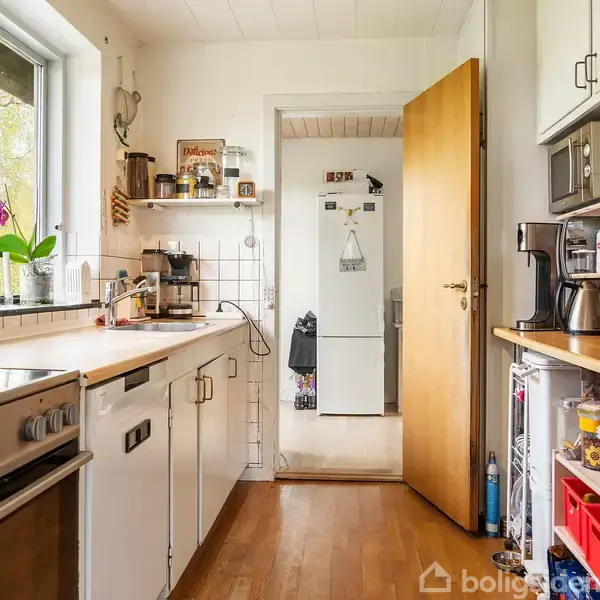 Grå sofa står vinklet i stuen, omgivet af et sofabord, en lænestol og en skænk med spiritus. Store vinduer åbner op til en solrig have med et dansk flag.