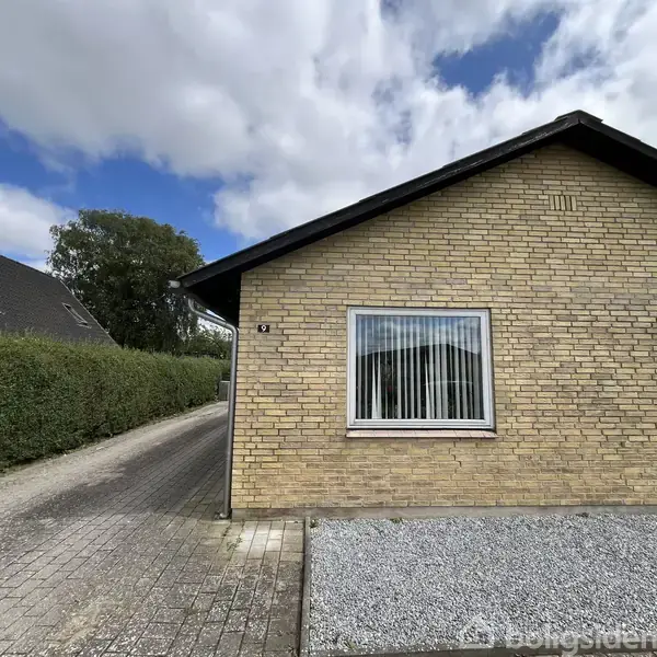 En gul murstensbygning med et vindue til højre. En flisebelagt sti løber langs siden omgivet af hække, under en skyet himmel. Husnummeret 9 er synligt ved døren.