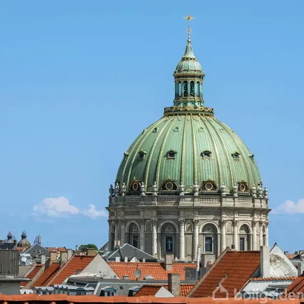 En stor, grøn kuppel troner over byens tage. Den arkitektoniske struktur er omgivet af røde teglstenstage under en klar blå himmel, hvilket skaber en historisk bymæssig atmosfære.