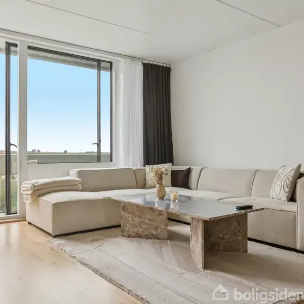 En beige hjørnesofa står i en lys stue ved siden af en stor glasdør, som giver udsigt til en balkon. Et marmorbord står på et tæppe foran sofaen.