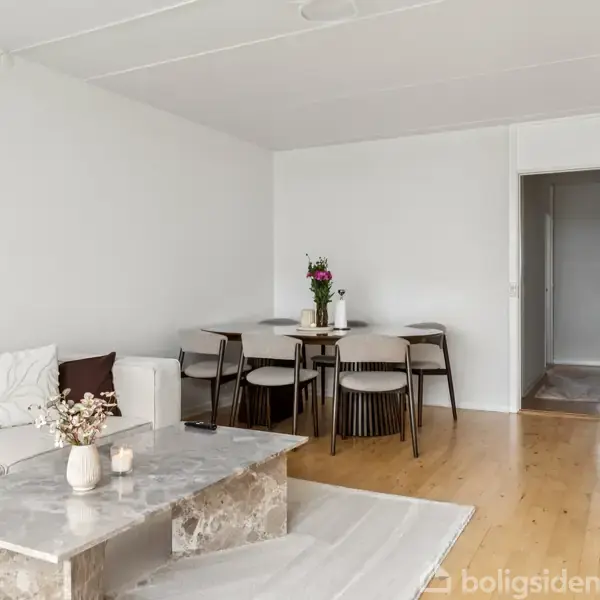 En hvid stue indeholder en sofa, et marmorbord med dekorationer og en spisebordgruppe med stole. Rummet er minimalistisk indrettet med lyse trægulve og hvide vægge.