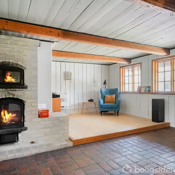 En hvid pejseovn med ild i brænder roligt. Det står i et rum med træbjælker i loftet, et blåt lænestol med pude og flere vinduer med naturligt lys.