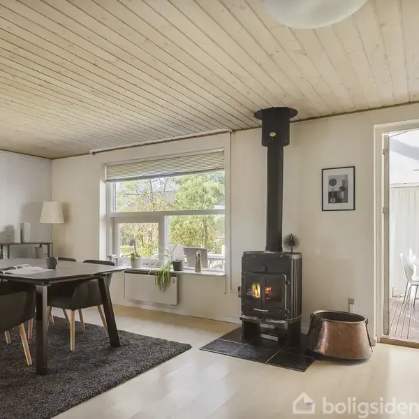 En brændeovn står i hjørnet af en stue, brænder og afgiver varme. Rummet har trægulv og træloft, spisebord med sorte stole og store vinduer, der giver udsyn til en terrasse.