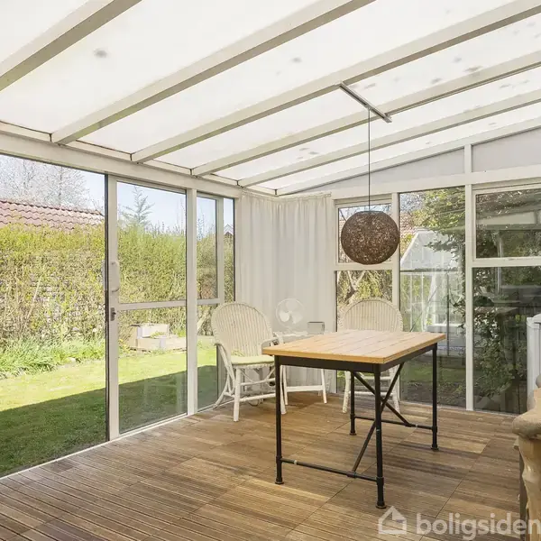 Et lyst udestueområde med træbord og stole, omgivet af glasdøre og gardiner. Udsigt til en grøn have udenfor. En rund lampe hænger over bordet.