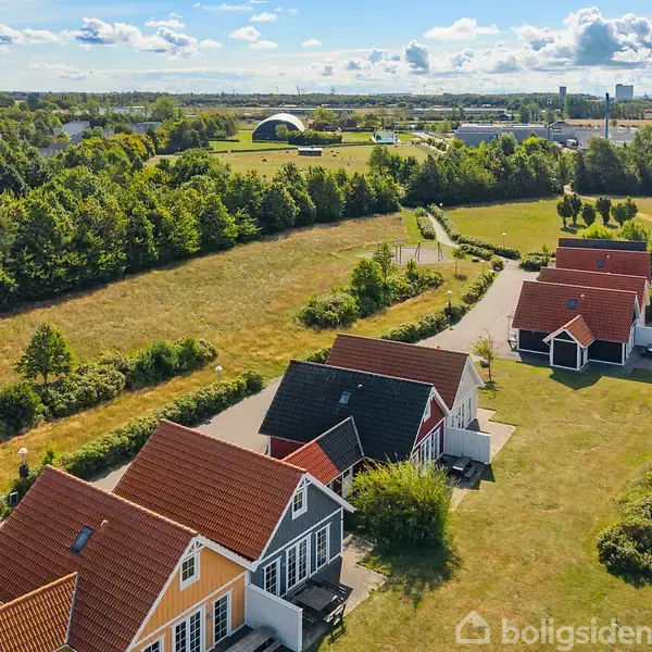 Farverige huse med røde tage står i række langs vej omgivet af grønne områder. Baggrund viser åbne marker og spredt beplantning under en skyet himmel.