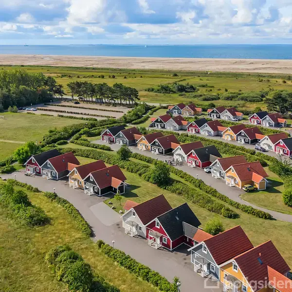Række af farverige huse ligger i et grønt område nær kysten, omgivet af stier og åbne græsarealer, med havet og en skib i baggrunden.
