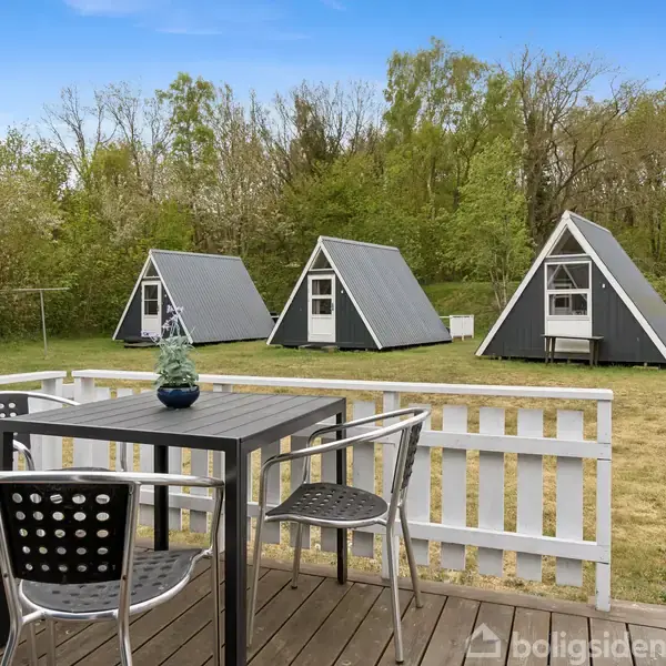 Terrassebord med stole står på træveranda. I baggrunden er tre A-formede, grå træhytter på græsplæne. Træbevoksede omgivelser skaber en naturlig ramme.