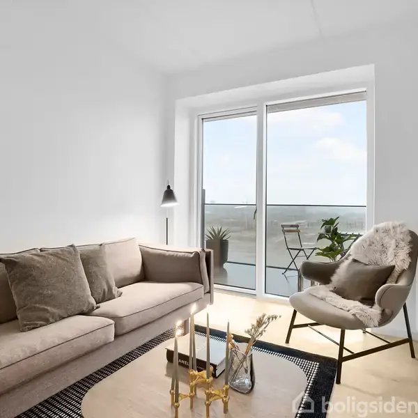 Sofaen står i en lys stue ved siden af en pude og gulvlampe, med udsigt til en altan med stole og planter gennem store vinduer.