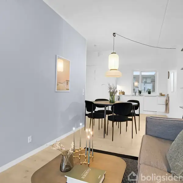 En moderne stue med grå sofa og sofabord, dekoreret med bøger og lys. I baggrunden ses spiseplads med sorte stole og køkken. Lyse vægge og hylder skaber et minimalistisk miljø.