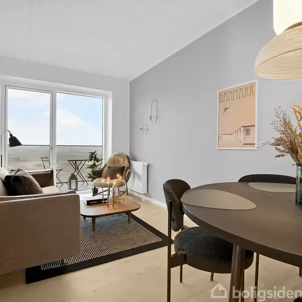 En moderne stue med en sofa ved et stort vindue. Spisebord med stole og tørrede blomster. Udendørs altan ses gennem døren, og der er minimalistisk kunst på væggen.