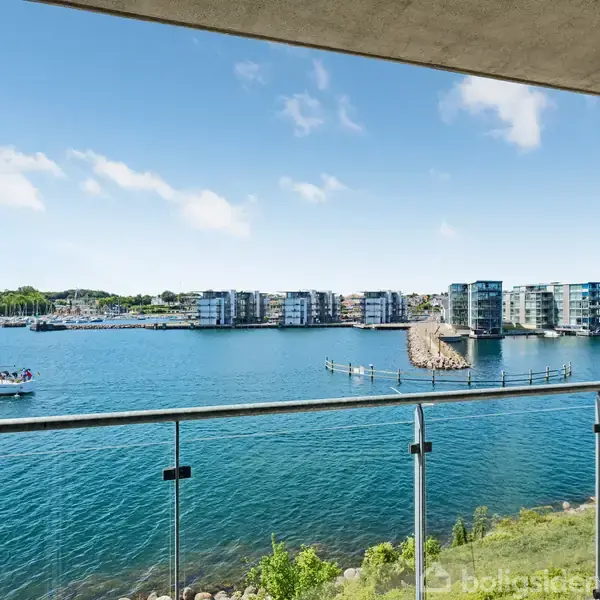 En glasbalustrade omkranser en altan med udsigt over en rolig blå fjord, hvor en sejlbåd driver forbi. I baggrunden ses moderne lejlighedsbygninger og grønne områder langs vandkanten.