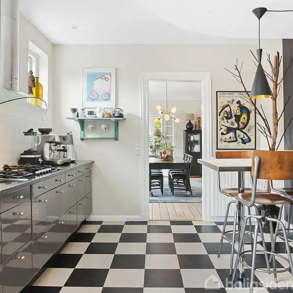 Køkken med skakternet gulv; moderne indretning med sort-hvide skabe, gaskomfur og emhætte. Barområde med høje stole. Vægkunst og dekorationer hænger ved spisebord i baggrunden.