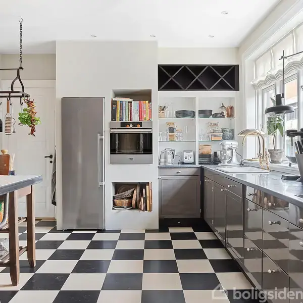 Et moderne køkken med sort-hvidt ternet gulv. Køkkenet har en lang bordplade med forskellige køkkenredskaber, en indbygget ovn, åbne hylder med bøger, og et stort køleskab ved siden af spiseområdet.