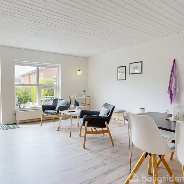En lys stue indeholder moderne stole og et bord, placeret mod en hvidmalet væg med billeder. Store vinduer lader naturligt lys strømme ind, med udsigt til en grøn have.