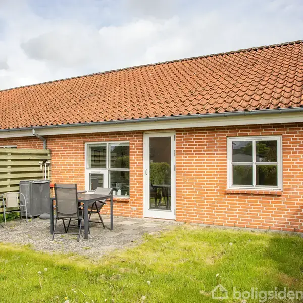 Rækkehus med rød murstensfacade og rødt tegltag. Foran står havemøbler på terrassefliser, omkranset af grøn græsplæne og hæk. Huset har vinduer og en havedør.