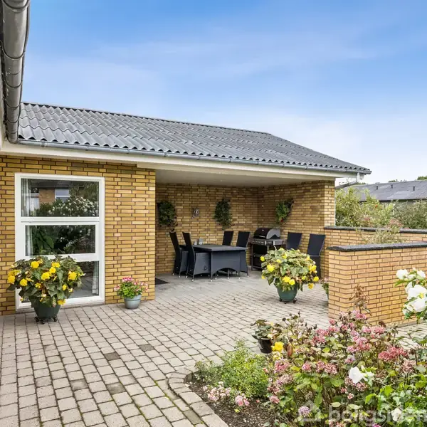 En udendørs terrasse med gule murstensvægge har en spiseplads med bord og stole. Omkring terrassen er der blomster og grønne planter. Himlen er blå og klar.