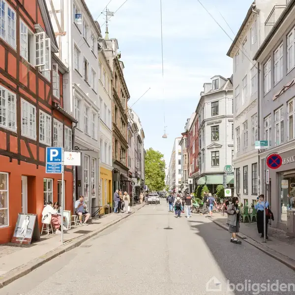 Gade med farverige bygninger på hver side, mennesker gående og cyklende. En parkeringstavle og caféborde ses i venstre side, meget livlig atmosfære på en solskinsdag.