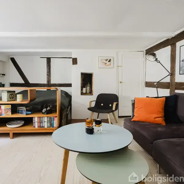 En moderne stue med en rund sofabord foran en mørk sofa med orange puder. En bogreol fungerer som rumdeler ved sengen. Lyse gulve og træbjælker skaber varme.