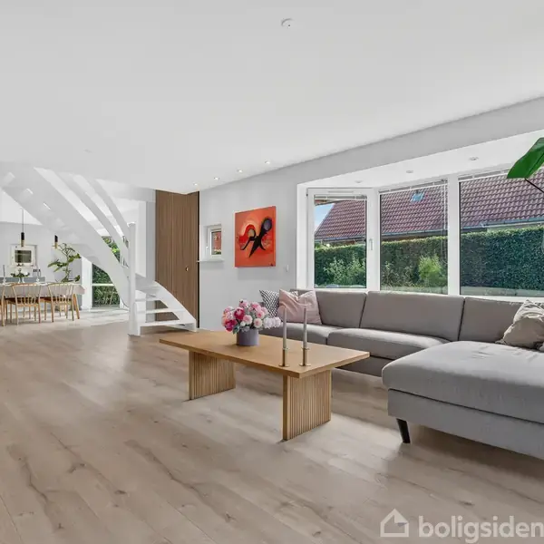 Stor, lys stue med grå hjørnesofa ved vindue. Træbord står foran sofaen. Trappen leder til en spisestue med træbord og stole. Planter og kunstværker dekorerer rummet.
