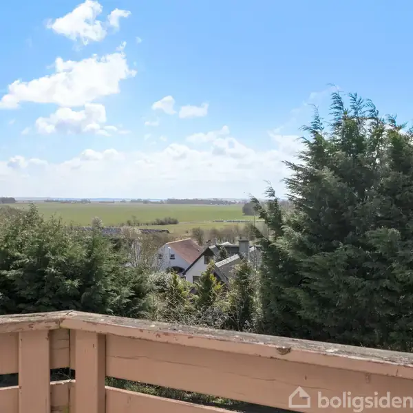Træhegn danner en terrassekant; i baggrunden ses grønne træer og åbne marker under en blå himmel med skyer.