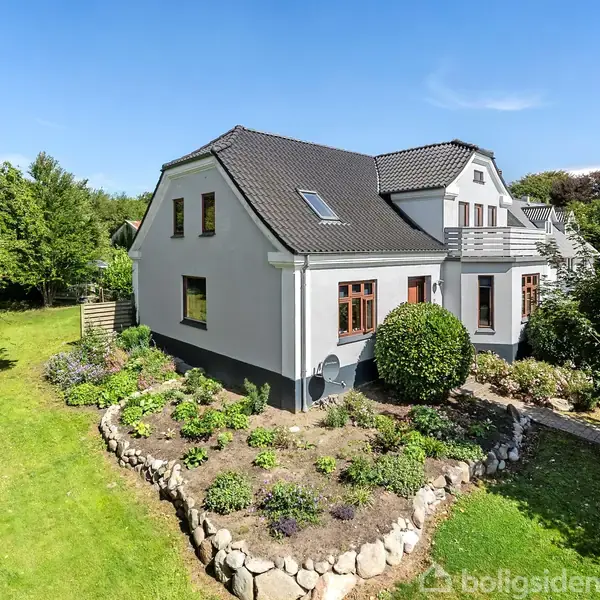 En hvid villa med sort tag står i en frodig have. Huset har flere vinduer og en altan. Haven er velplejet med græsplæne, træer og blomsterbede indrammet af sten.